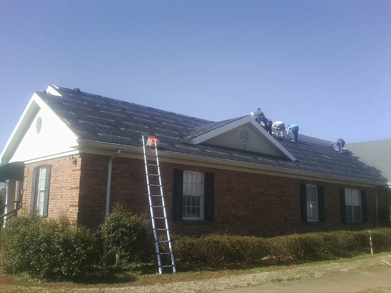 Crew tearing shingles off a brick ranch roof with a ladder set — Metal Roof Repair work by our crew in Milton Center