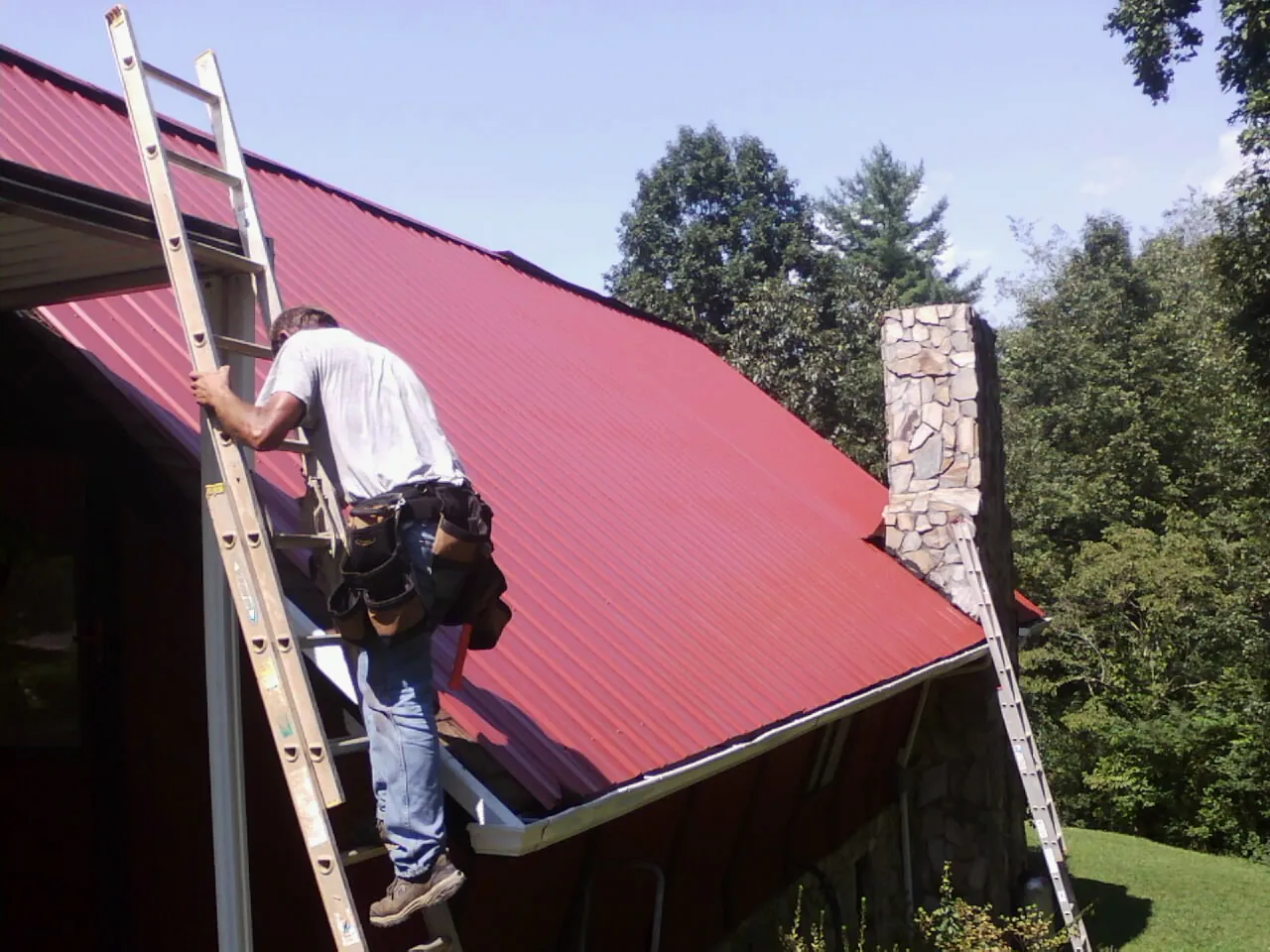 Roofer on a ladder beside a red metal roof and stone chimney — Soffit & Fascia Repair by Roofing Brothers in Milton Center